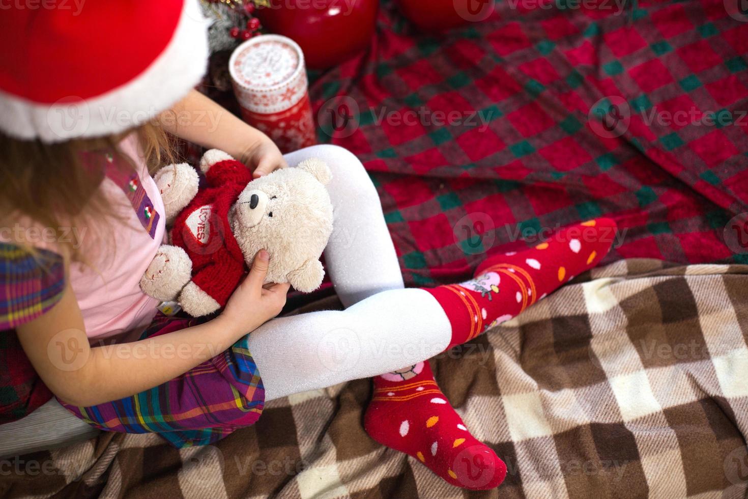 A little girl holding a Teddy bear, sitting on a plaid blanket in the Christmas decorations near a Christmas tree with boxes of gifts and a Santa hat. New year, children's game photo