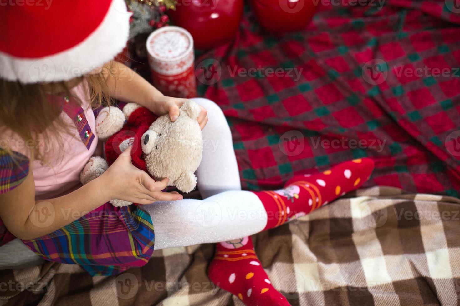 A little girl holding a Teddy bear, sitting on a plaid blanket in the Christmas decorations near a Christmas tree with boxes of gifts and a Santa hat. New year, children's game photo