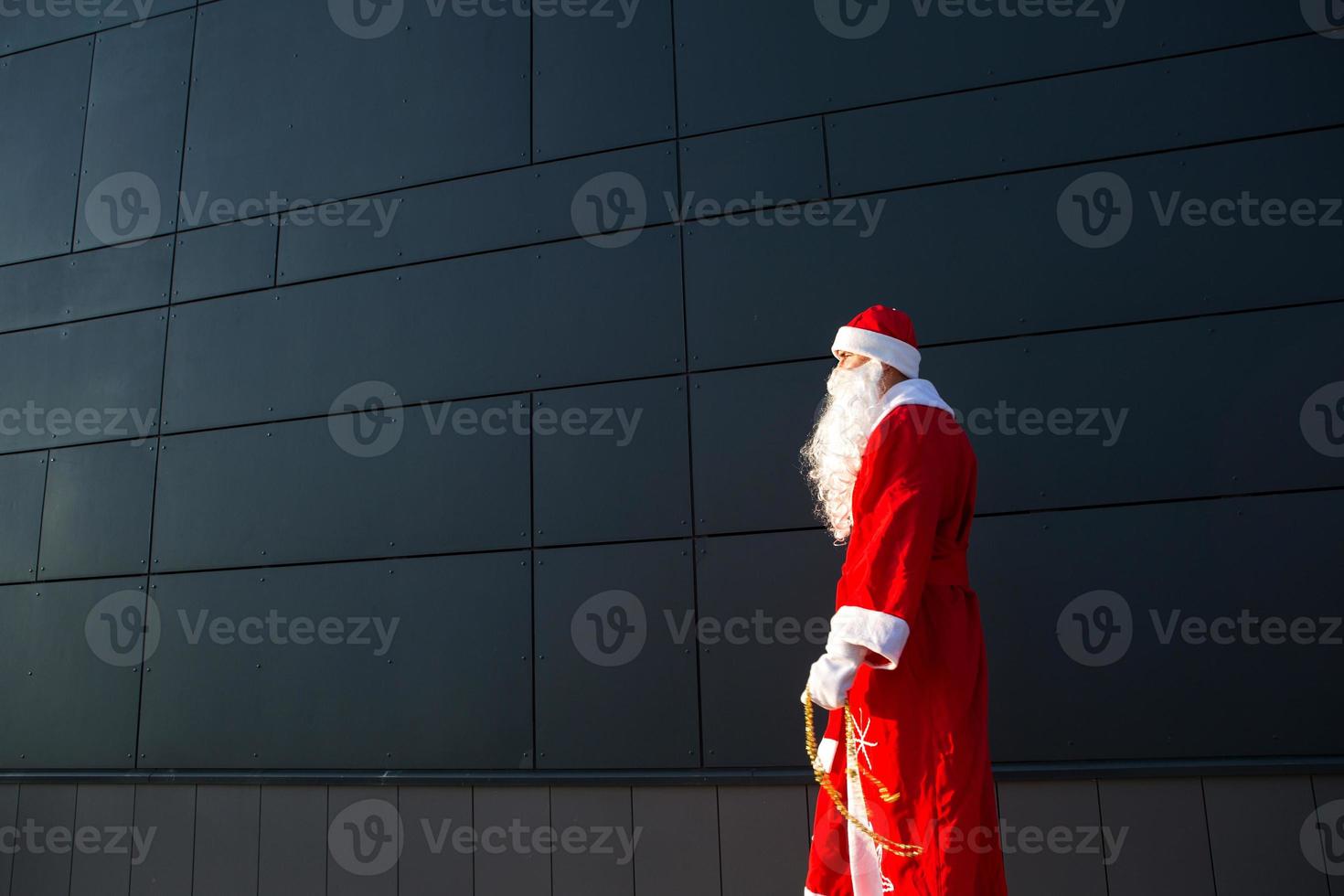 un joven vestido como santa en la calle, parado contra una pared moderna gris. barba blanca, traje rojo con sombrero. navidad, año nuevo. helada del abuelo ruso. fondo moderno gris, espacios de copia foto