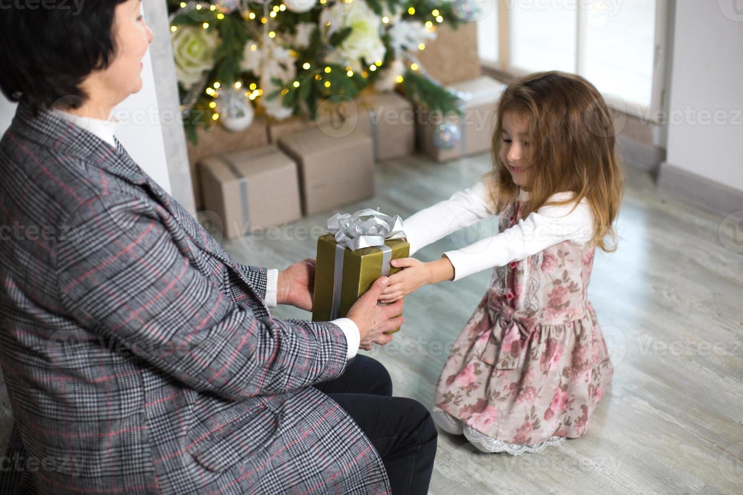 la abuela le da a su nieta un regalo para navidad. la niña extiende sus manos hacia la caja con el lazo. año nuevo, vacaciones familiares, comunicación de generaciones. foto