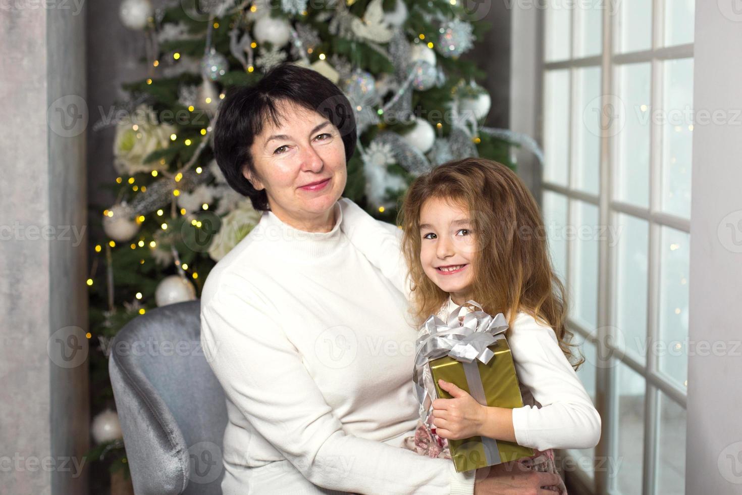 Grandmother with a little girl on the background of Christmas decorations and a large window. Family holiday, emotions, gift box. Granddaughter on granny's lap. New Year photo