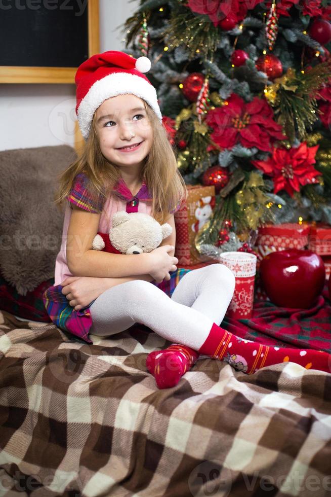 A little girl holding a Teddy bear, sitting on a plaid blanket in the Christmas decorations near a Christmas tree with boxes of gifts and a Santa hat. New year, children's game photo