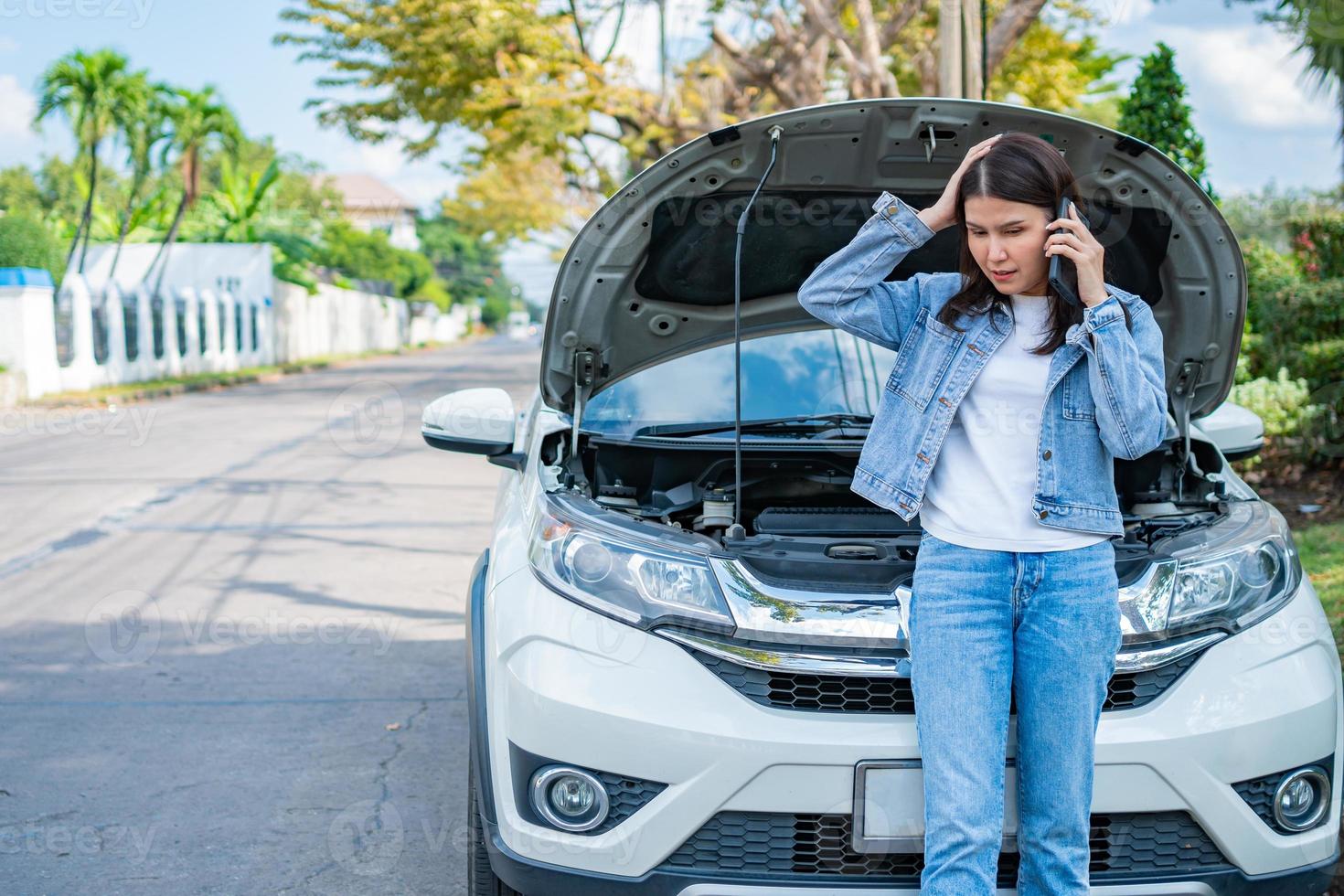 Angry Asian woman and using mobile phone calling for assistance after a car breakdown on street. Concept of vehicle engine problem or accident and emergency help from Professional mechanic photo