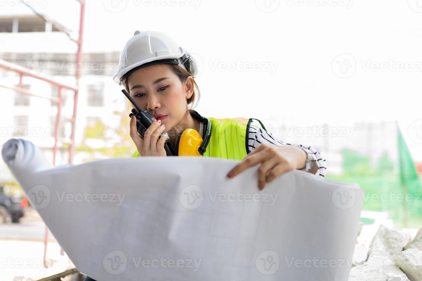 Asian engineer or Young woman Architect put on a helmet for safety and look at Blueprint for Inspect Building factory Construction Site and use walkie talkie for a talk with a contractor. photo