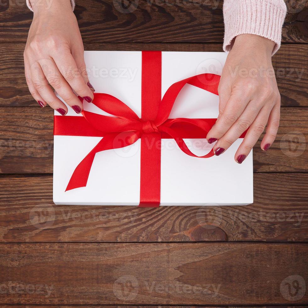 Woman's hands wrapping christmas, holiday handmade present in paper box with red ribbon. Give present box, decoration of gift on vintage wooden table, top view, copy space mock up template for design. photo