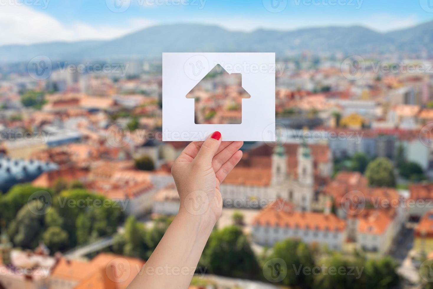 Woman's hand is holding a paper with a house cut out on it, panorama of the city in background. The concept of renting or buying real estate photo
