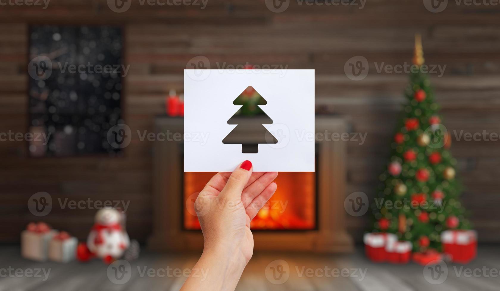 la mano sostiene un papel con un árbol de navidad recortado. chimenea con fuego, un árbol de navidad y adornos navideños foto