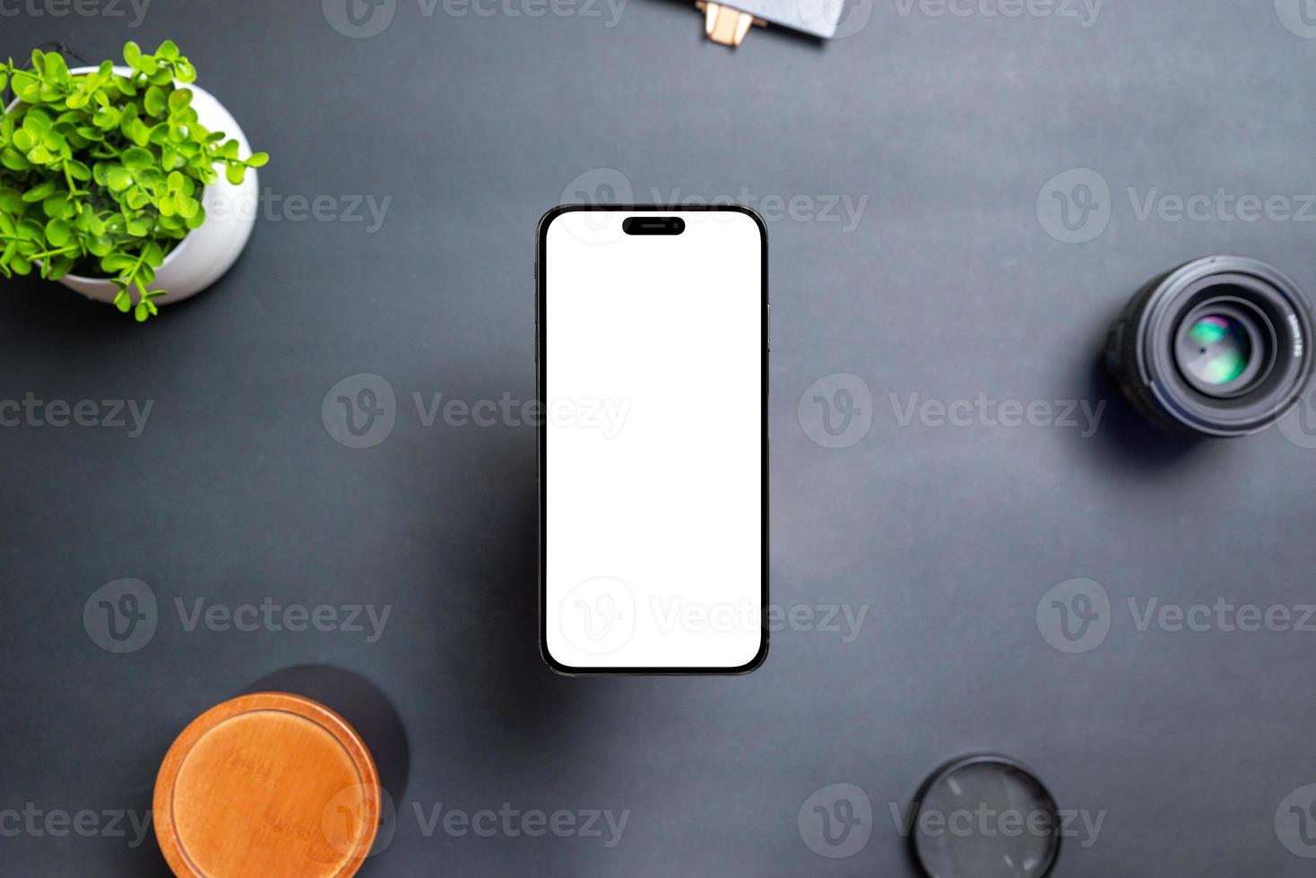 Phone with isolated display floats above the desk. The front camera is built into the display. Modern round and thin edges. Black table with plant, lens and box. Top view, flat lay photo