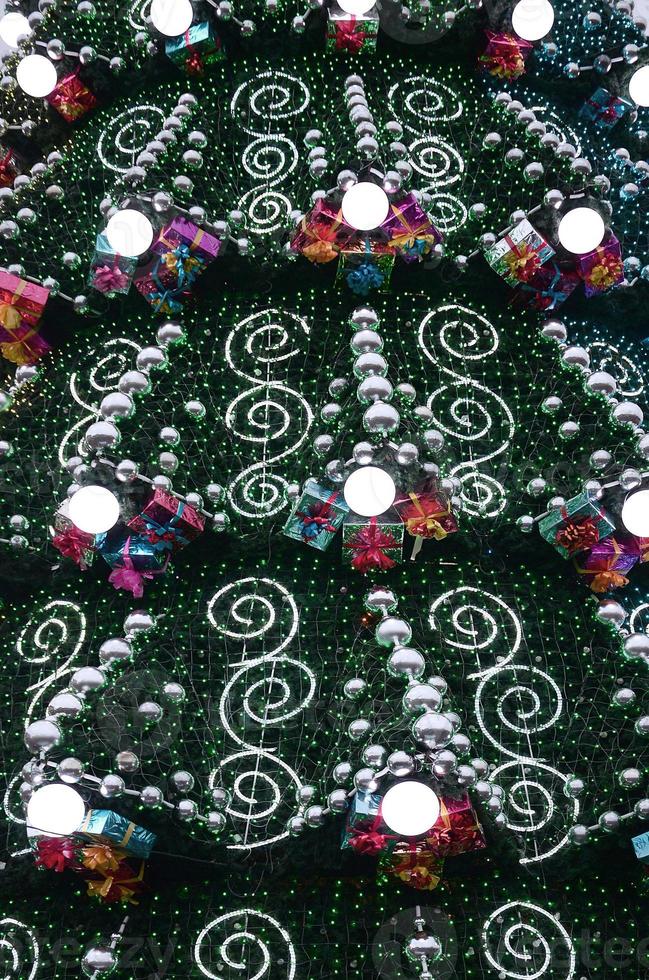 A fragment of a huge Christmas tree with many ornaments, gift boxes and luminous lamps. Photo of a decorated Christmas tree close-up