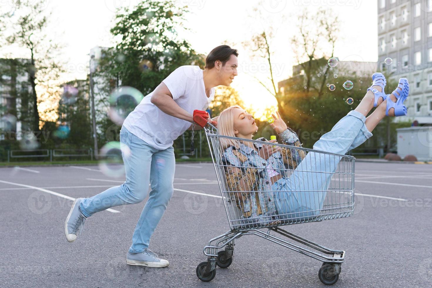 una pareja joven se divierte con un carrito de compras en el estacionamiento de un supermercado foto