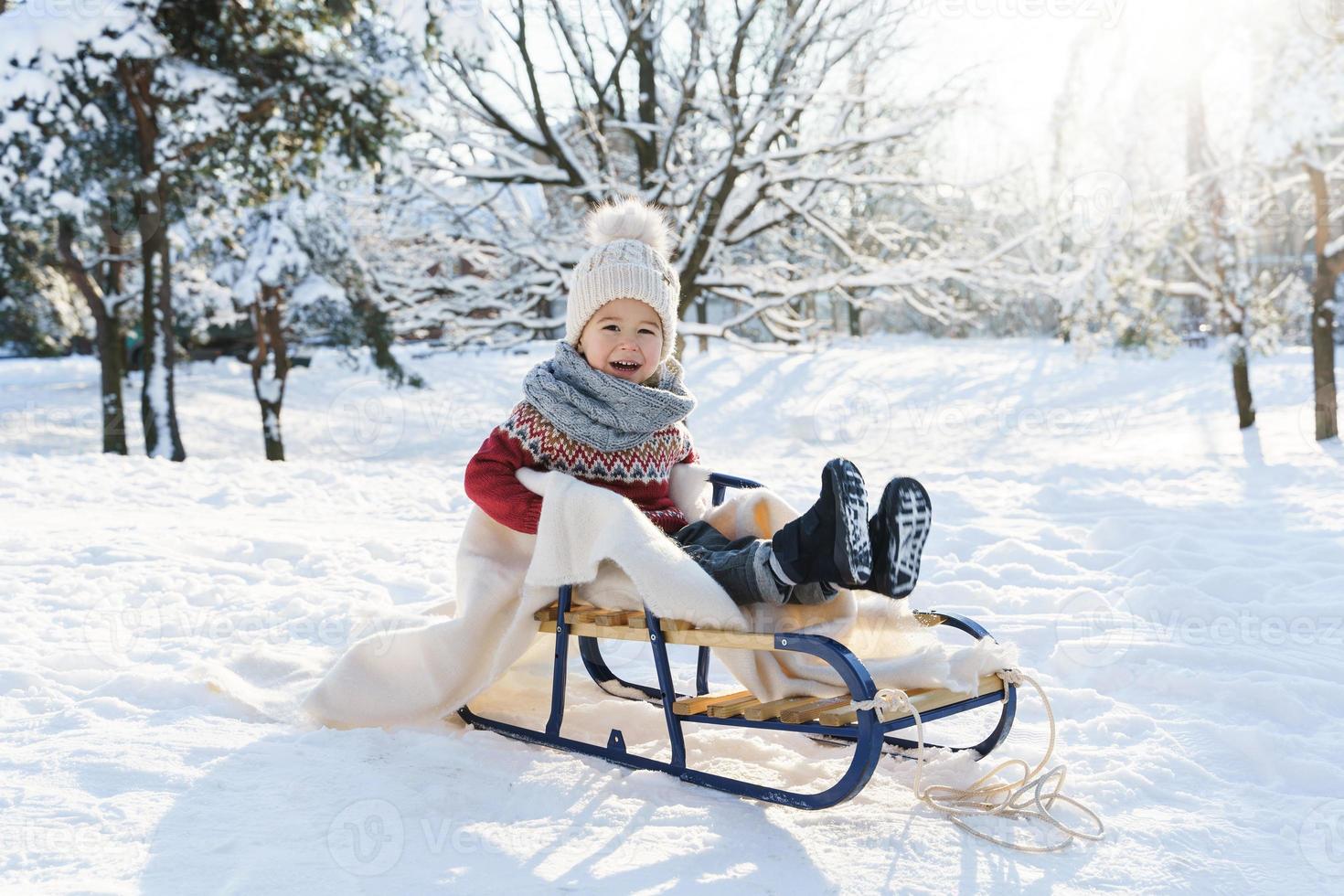 Toddler boy sitting on the sleigh in a snowy city park during sunny winter day photo