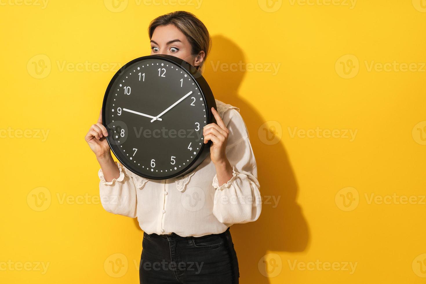 Businesswoman peeking out from behind big clock on a yellow background photo