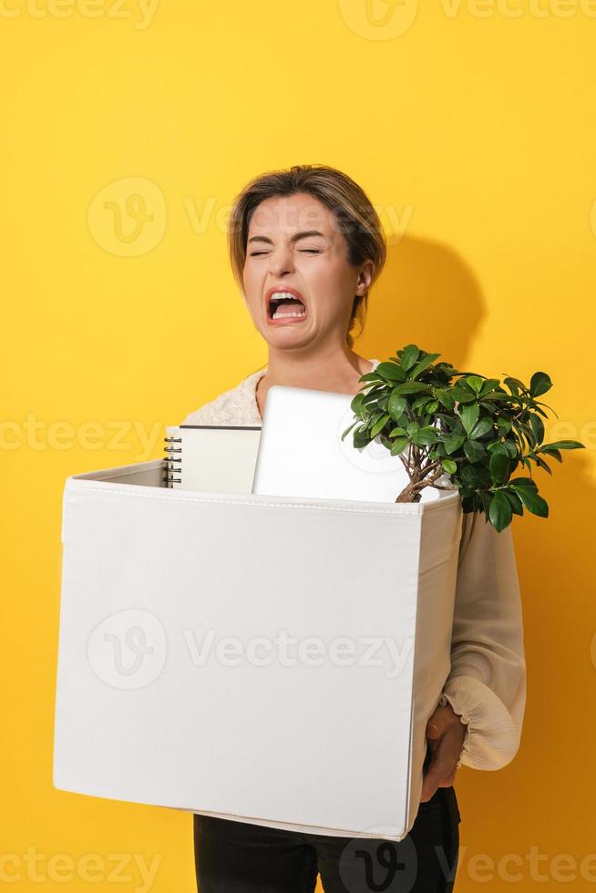 Upset woman holding box with personal items after job resignation against yellow background photo