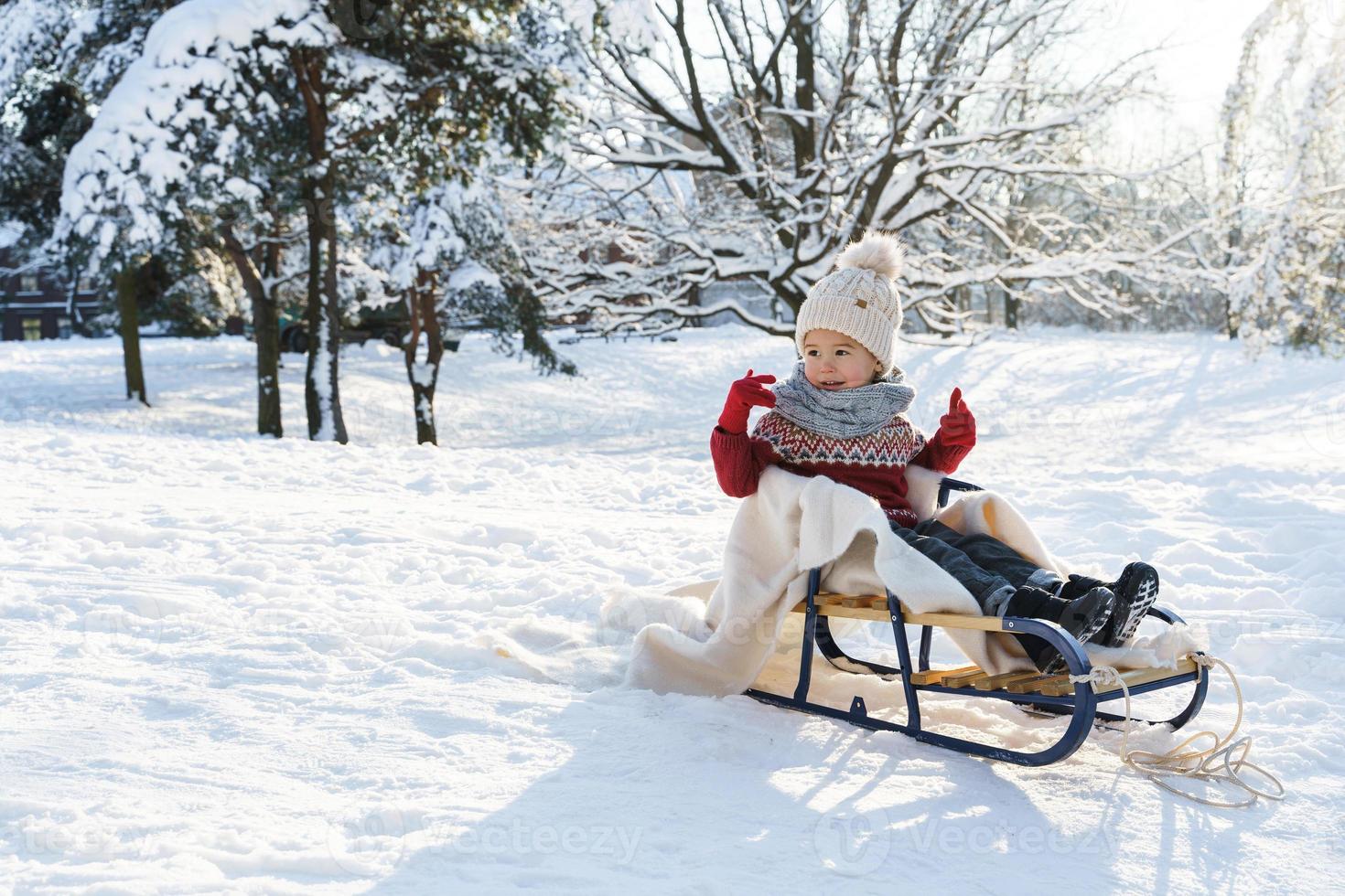 Toddler boy sitting on the sleigh in a snowy city park during sunny winter day photo