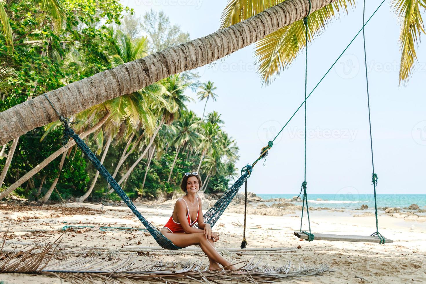 Young woman wearing red bikini relaxing in the hammock on sandy beach with coconut palm trees photo