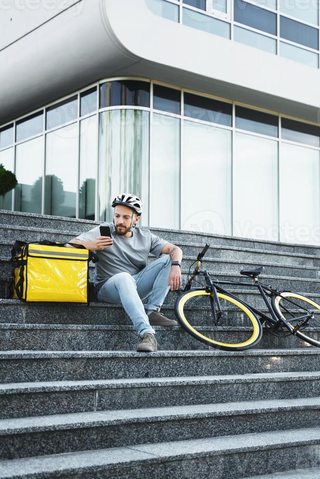 Express food delivery courier sitting on the stairs with insulated bag and bicycle. photo