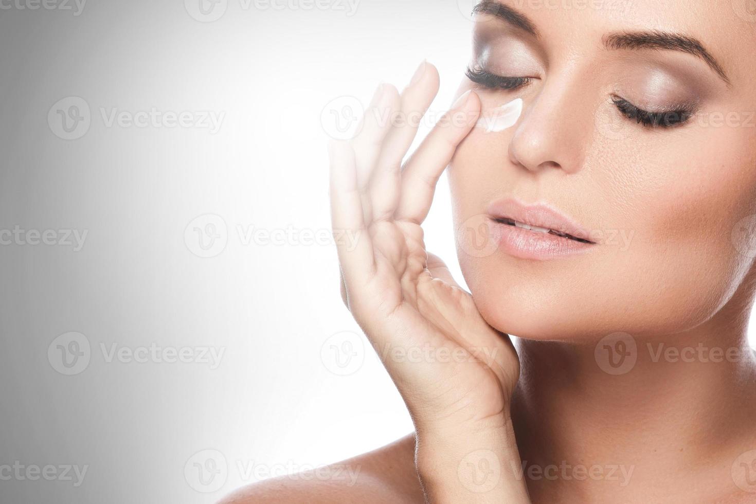 Beautiful woman applying moisturizing cream under her eye photo