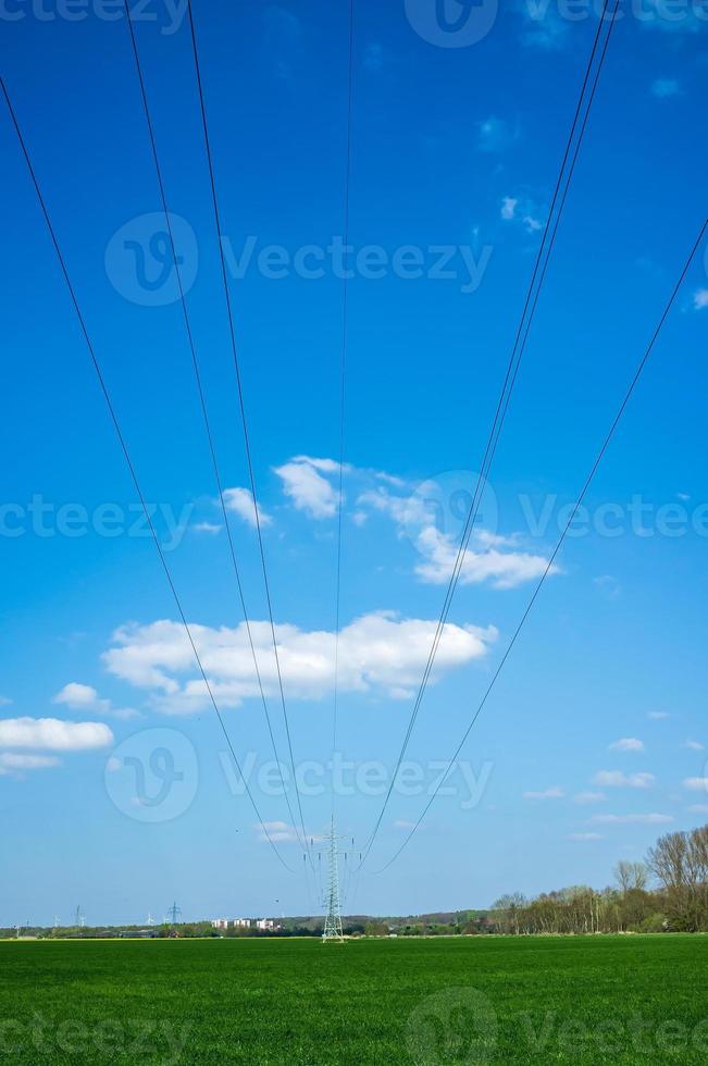 High metal electricity pylons with power lines that pass over an agricultural field, against the backdrop of a blue sky and clouds. photo