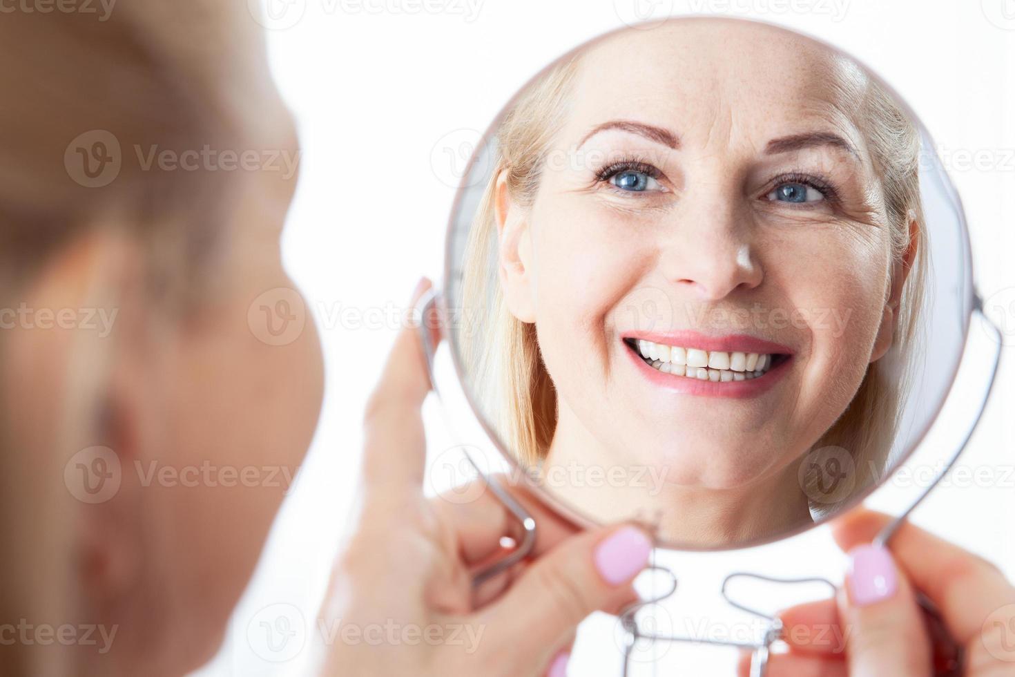 Happy middle aged woman model touching face skin looking in mirror. Smiling mature older lady enjoying healthy skin care, aging beauty, skincare treatment cosmetic products concept. photo