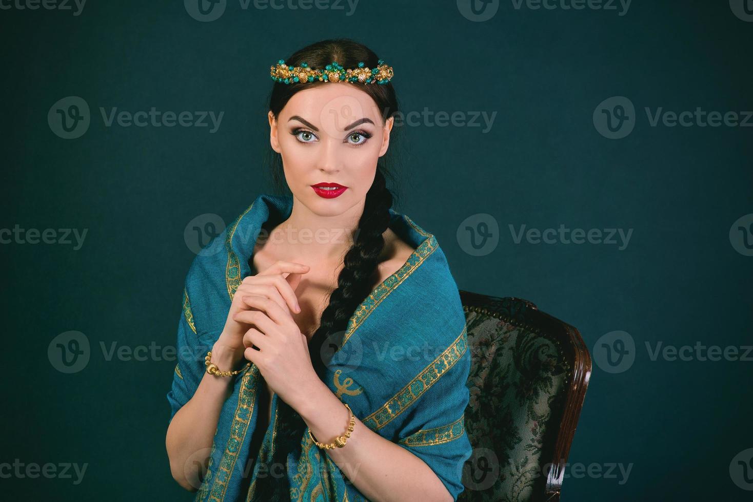 Portrait of young beautiful brunette in Indian style with wreath and braid in a blue scarf and gold bracelets on dark background photo