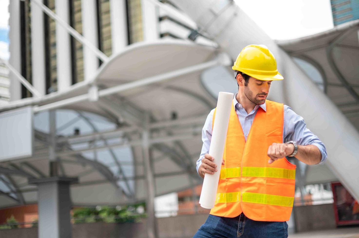 joven caucásico sosteniendo un papel grande, tipo con camisa azul claro y jeans con chaleco naranja y casco amarillo para seguridad en el área de construcción, mientras mira su reloj de mano. foto