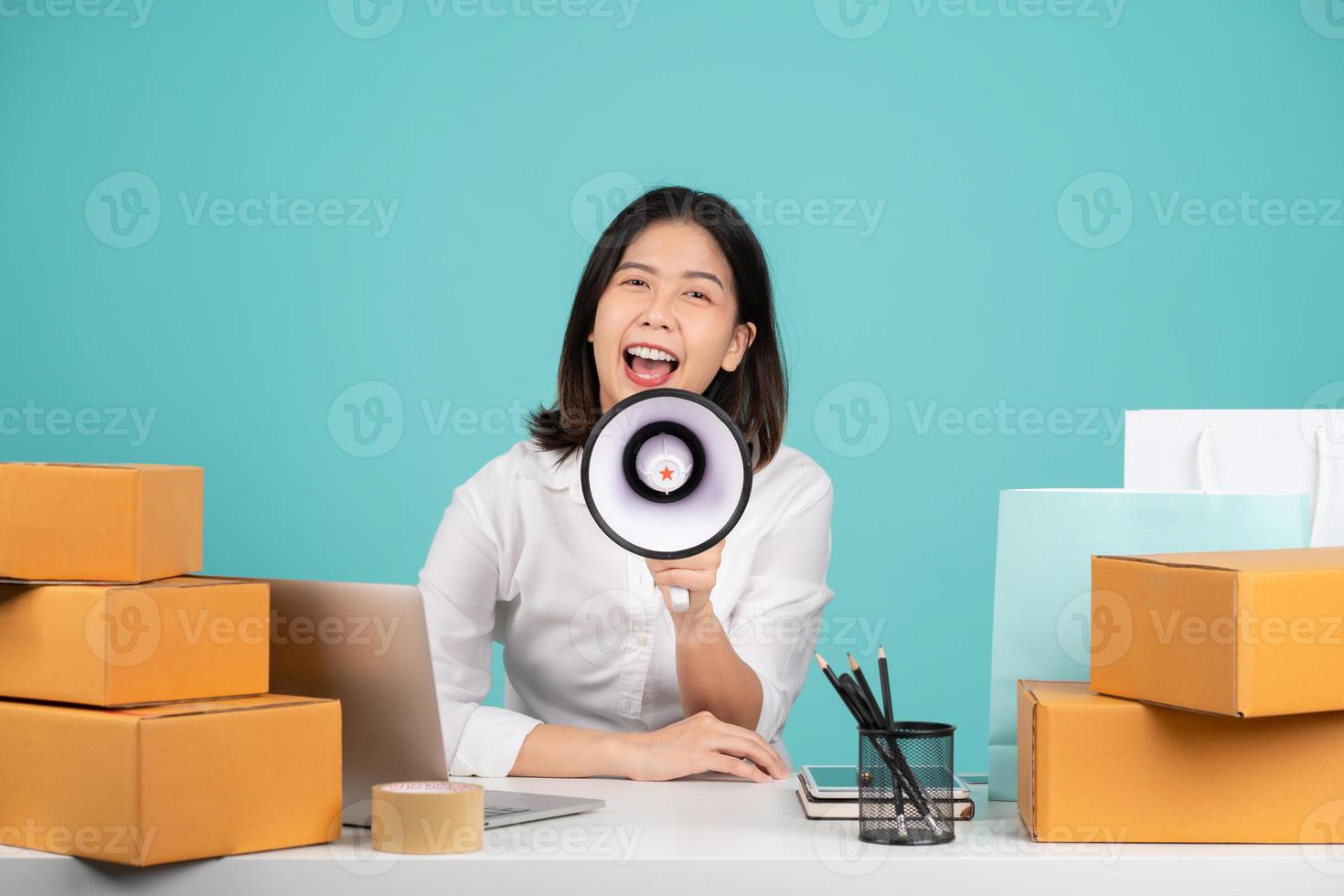 Asian businesswoman sitting at her desk with laptop and brown cardboard isolated on pastel green background. She was screaming and shouting with a megaphone in her hand. photo