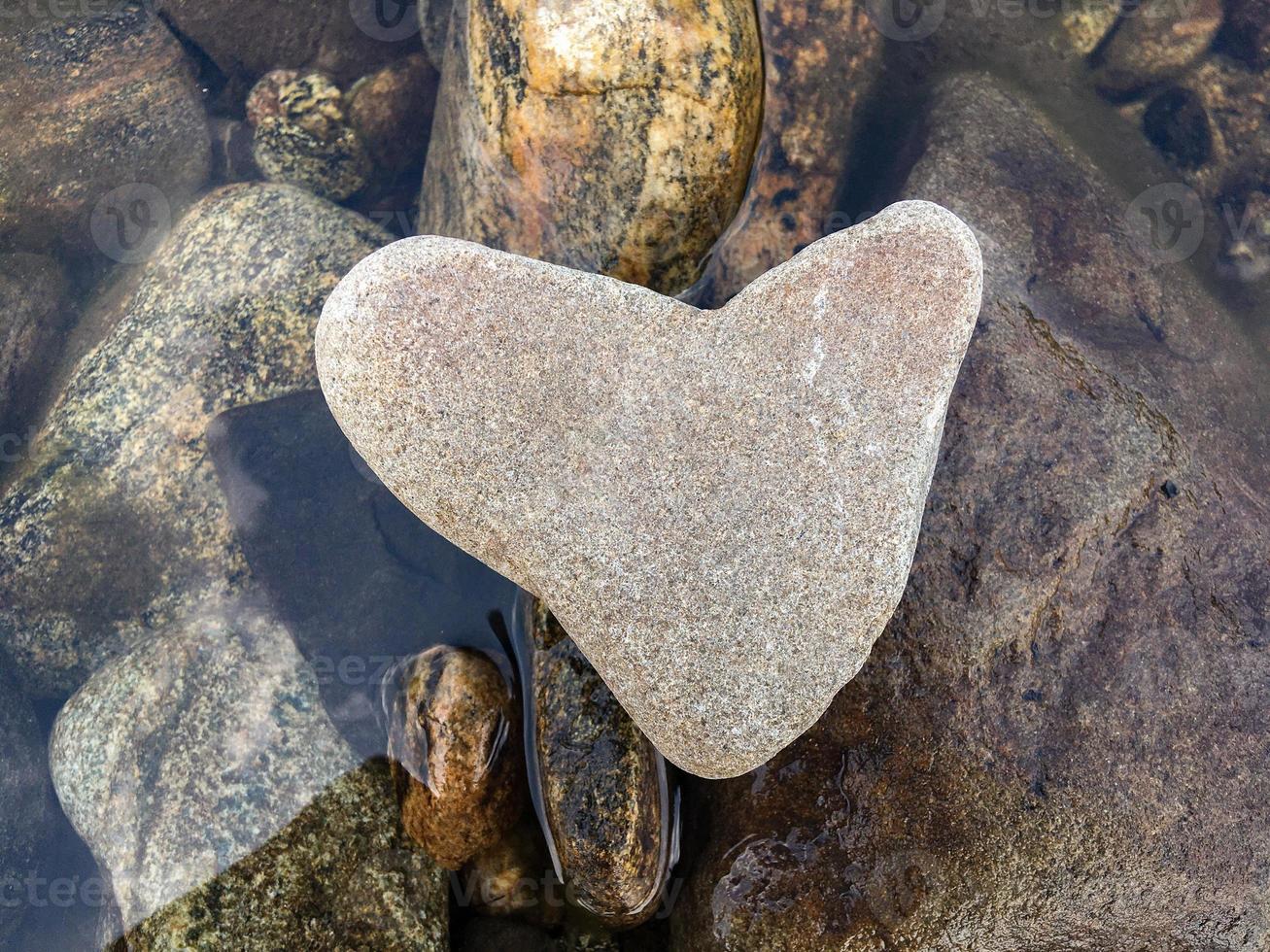 A heart-shaped stone lies on stones above the water, horizontal orientation photo