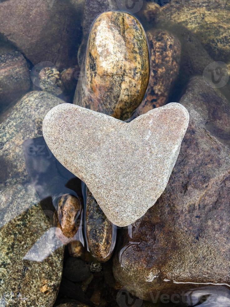 A heart-shaped stone lies on stones above the water, vertical orientation photo