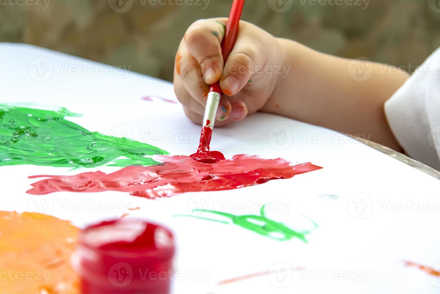 la mano de un niño sostiene un pincel y pinta papel con grandes trazos. manchas de pintura brillante sobre papel blanco. foto