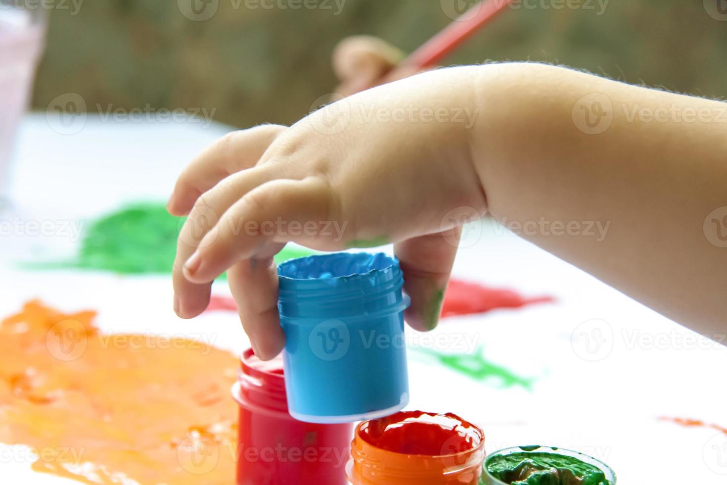 A child's hand holds a jar of paint about to draw with it. Smears of bright paint on white paper. photo