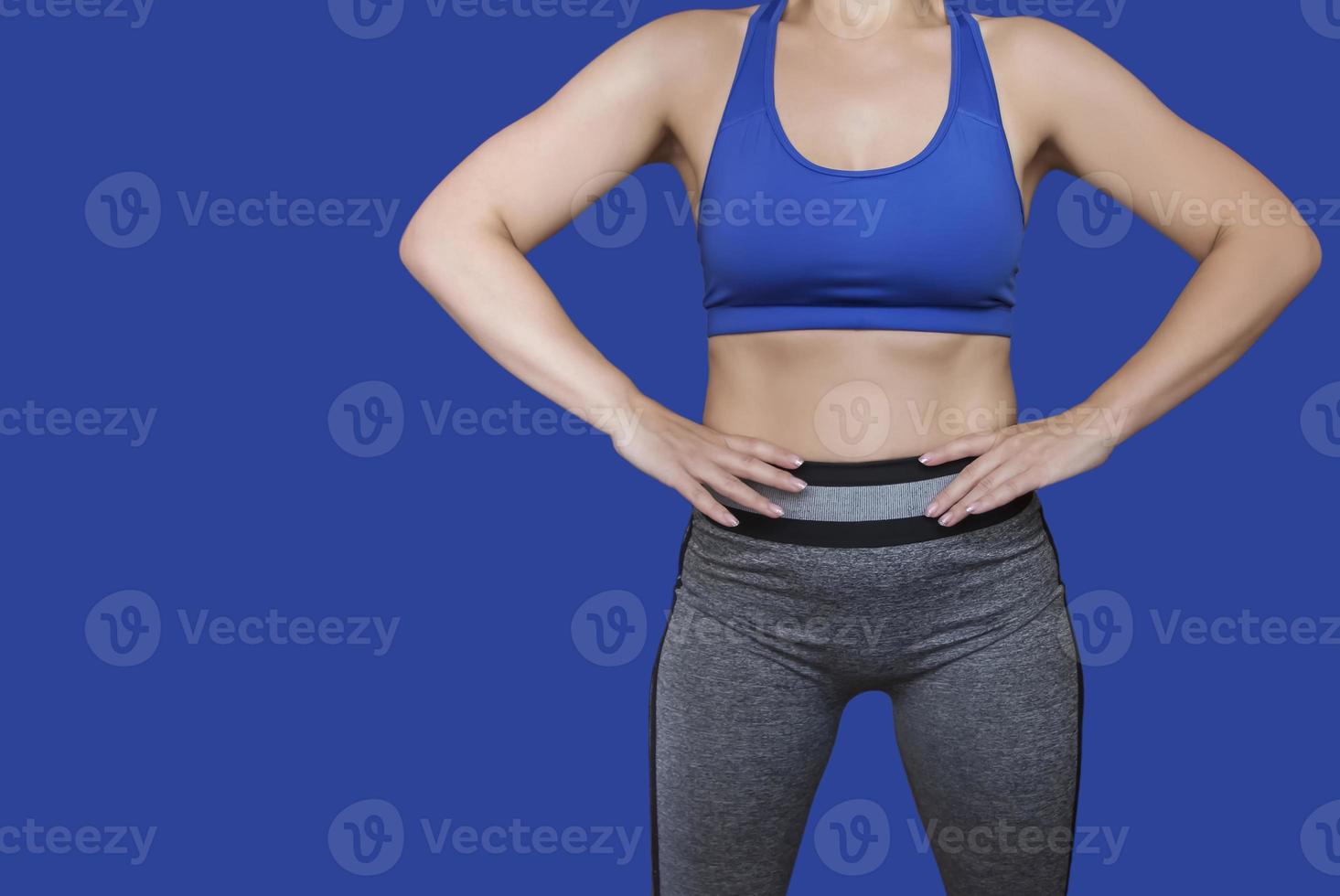 Fitness girl in a top and leggings points to her athletic figure. The Concept Of Weight Loss. Model on a blue background. A slender Young Lady Holds her hands at her waist. photo
