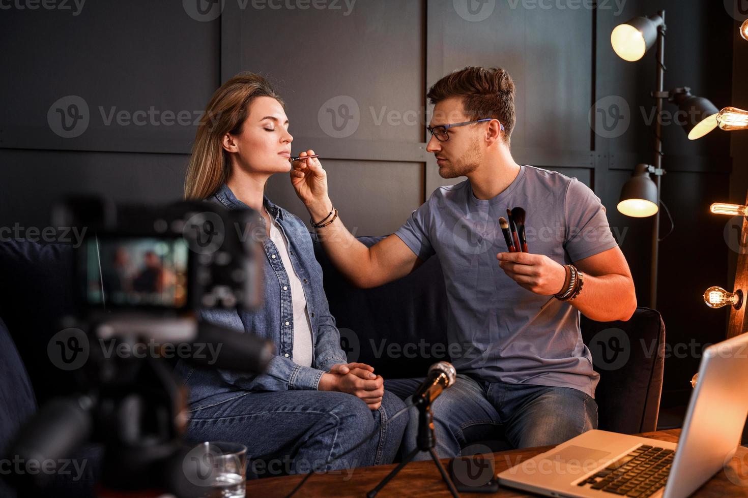 Young male beauty blogger is applying makeup to beautiful young woman photo