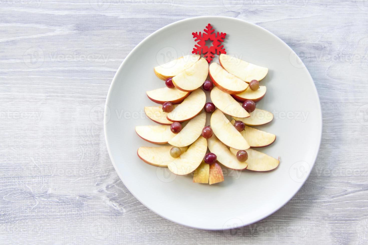 Christmas tree made of sliced apples and berries with a large red wooden snowflake instead of a star. Plate with apples on a light wooden background. photo