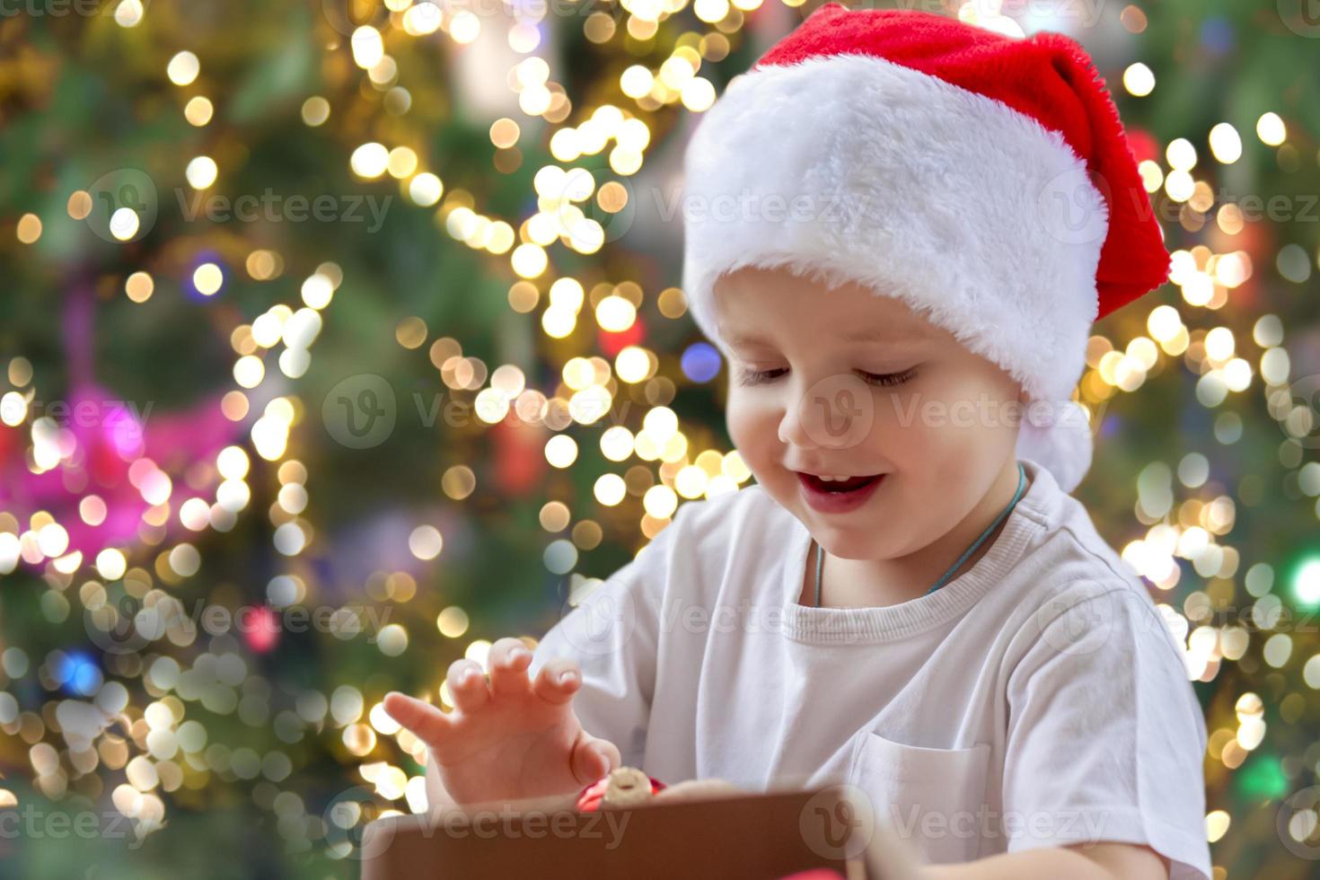 Child boy takes out of the box Christmas toys to dress up the tree. The Christmas tree is lit up and the baby looks at the Christmas toys with pleasure and joyful emotions. photo