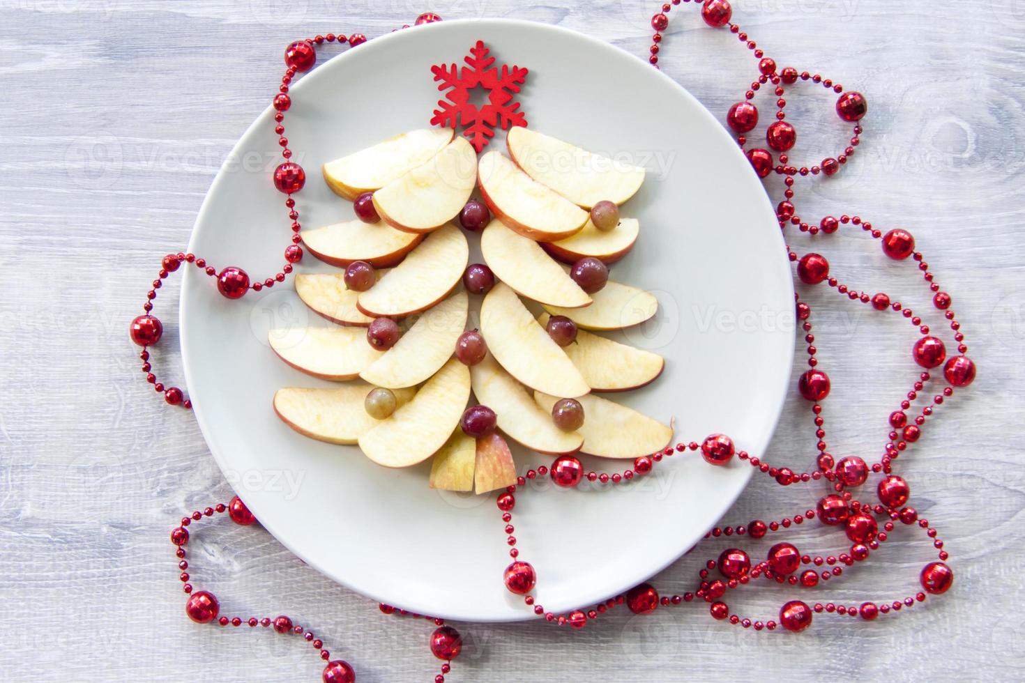 Christmas tree made of sliced apples and berries with a large red wooden snowflake instead of a star. Plate with apples on a light wooden background. photo