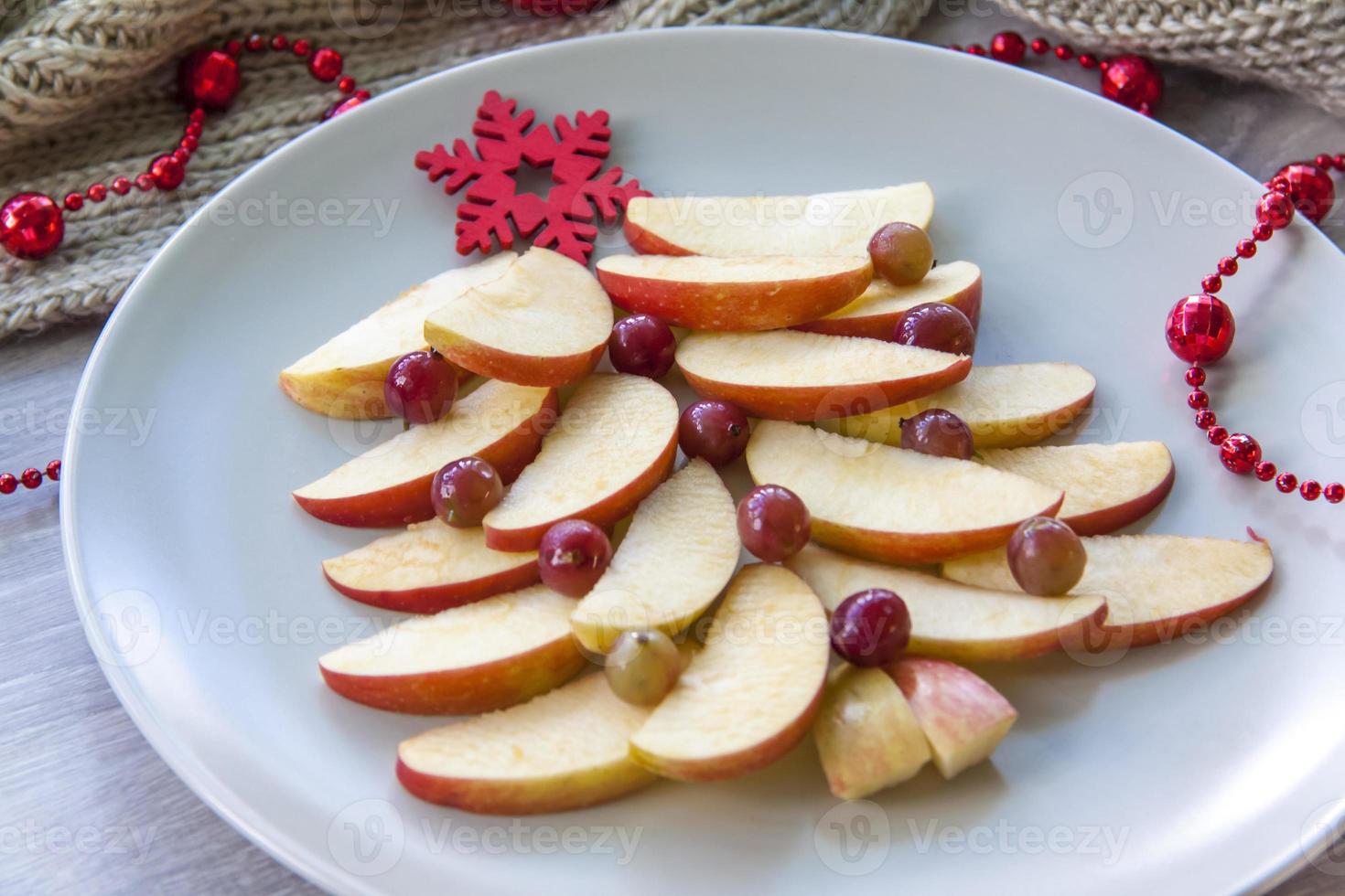 Christmas tree made of sliced apples and berries with a large red wooden snowflake instead of a star. Plate with apples on a light background. photo