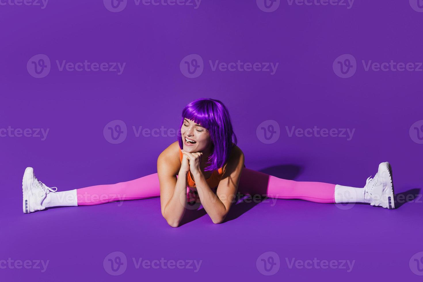 Cheerful woman wearing colorful sportswear doing splits against purple background photo