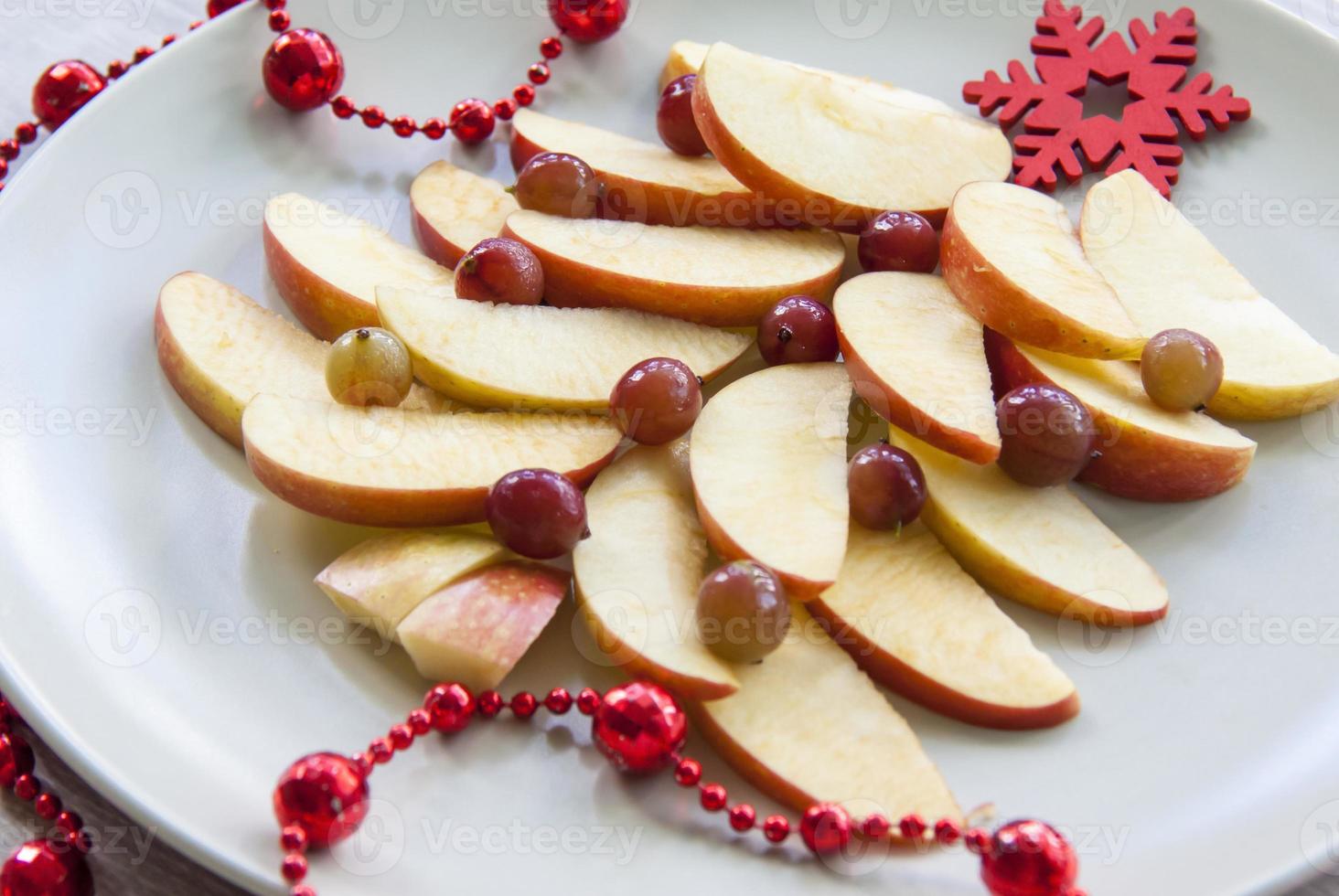 Christmas tree made of sliced apples and berries with a large red wooden snowflake instead of a star. Plate with apples on a light background. photo