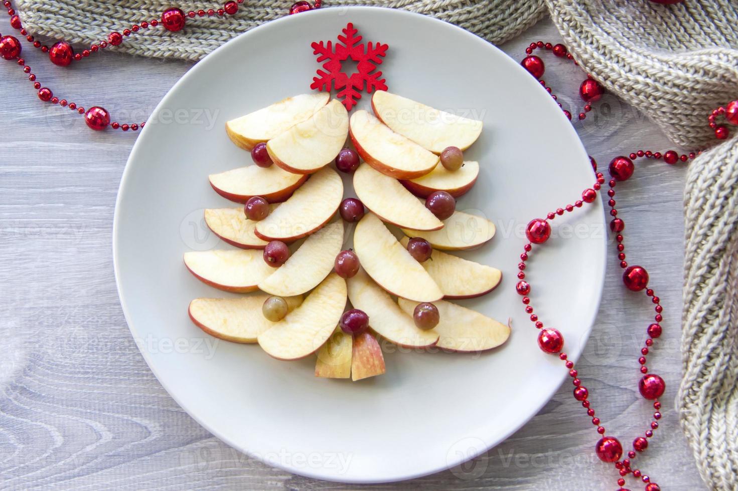 Christmas tree made of sliced apples and berries with a large red wooden snowflake instead of a star. Plate with apples on a light background. photo