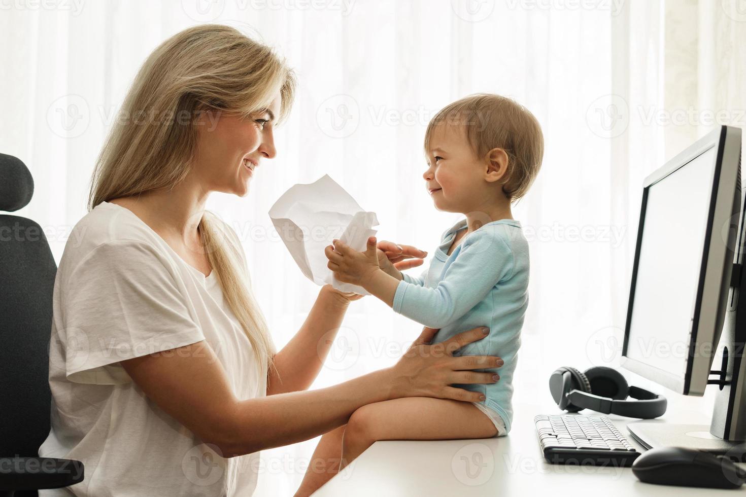 Little boy sitting on a desk distracting freelancer mother from paperwork. photo
