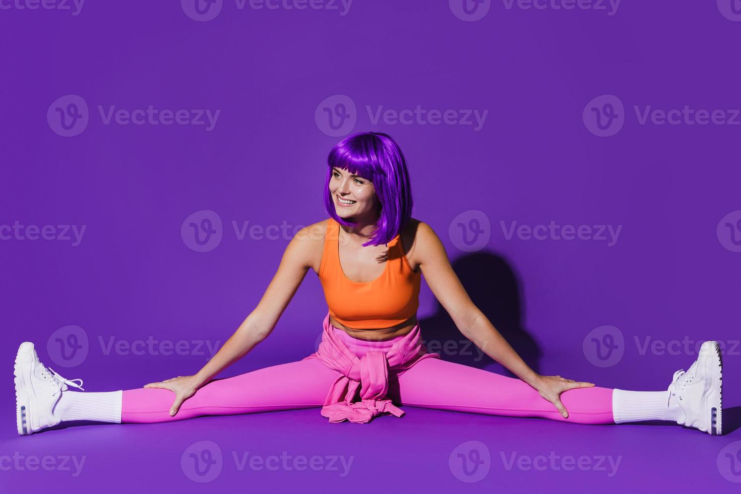 Cheerful woman wearing colorful sportswear doing splits against purple background photo