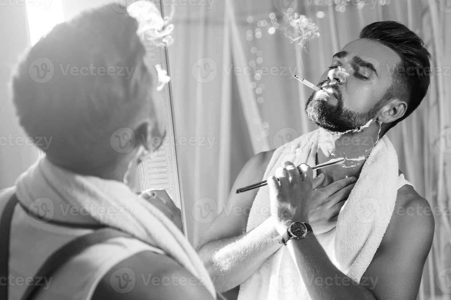Man looking in the mirror, smoking a cigarette and shaving his beard with a straight razor photo