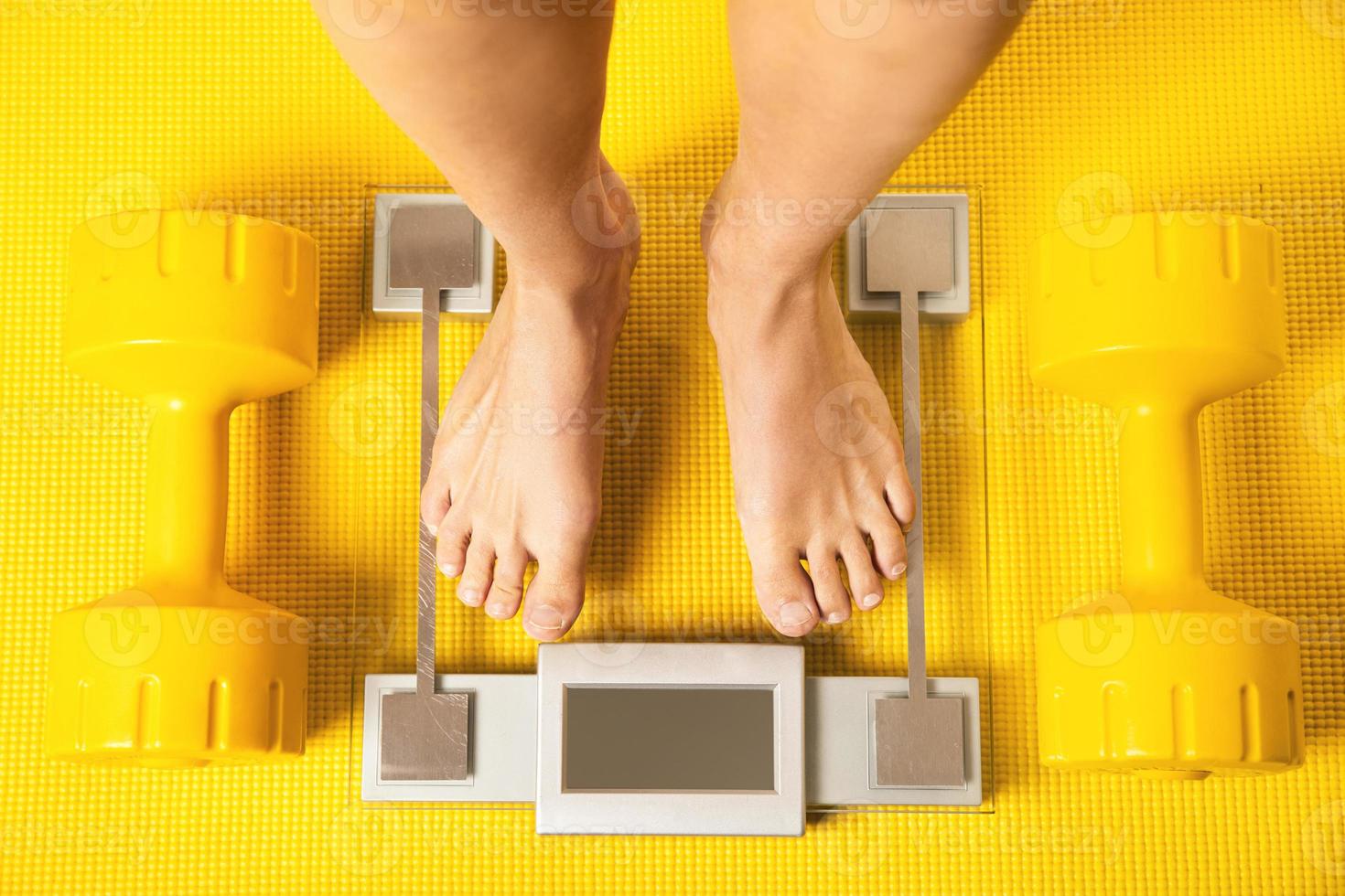 Female feet on the modern weighing scale with a yellow dumbbells and fitness mat photo