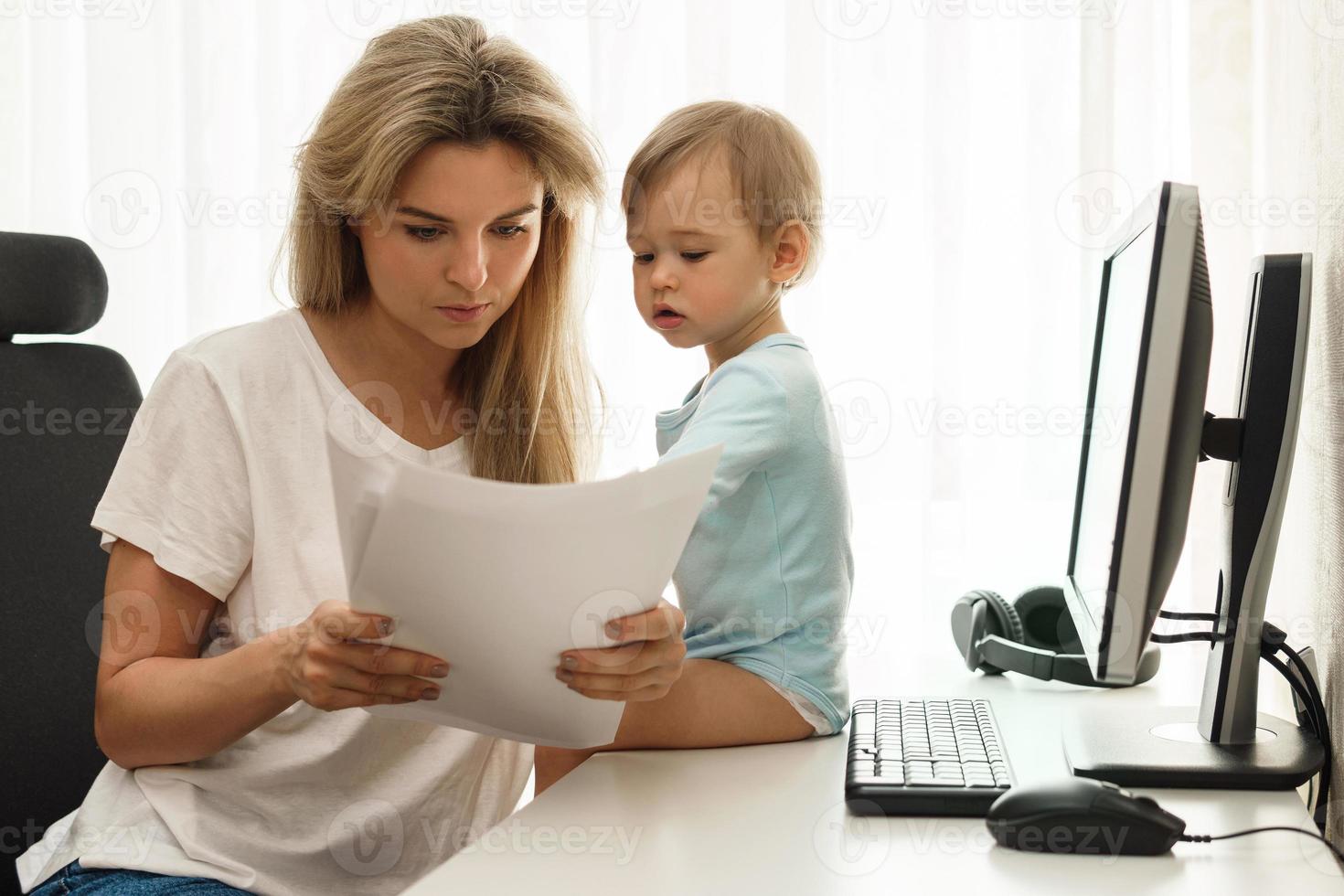 joven madre independiente leyendo documentos en la oficina de su casa con su hijo sentado en el escritorio. foto