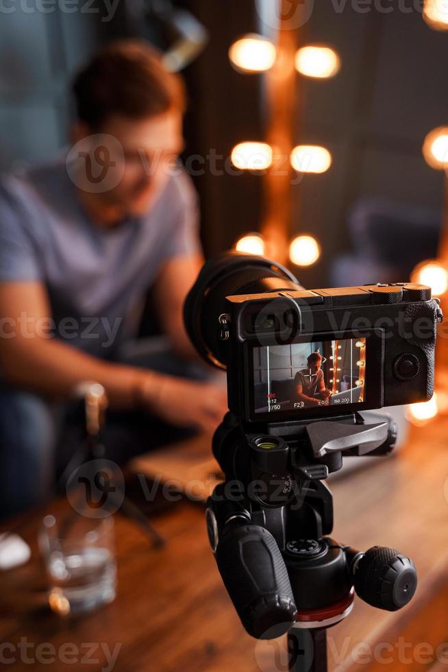 Blogger sitting on sofa in front of a camera is working on his laptop photo