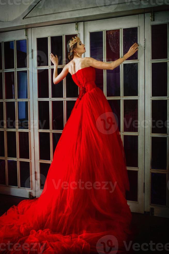 Beautiful woman in long red dress and in royal crown standing near retro fireplace door with stained-glass windows photo