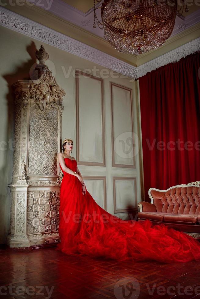 Beautiful woman in long red dress and in royal crown near fireplace in luxury interior in photo studio