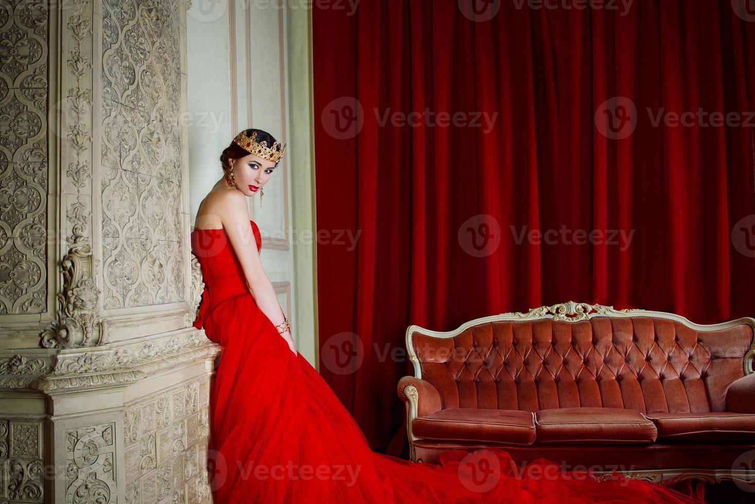 hermosa mujer con vestido rojo largo y corona real cerca de la chimenea en interiores de lujo en estudio fotográfico foto