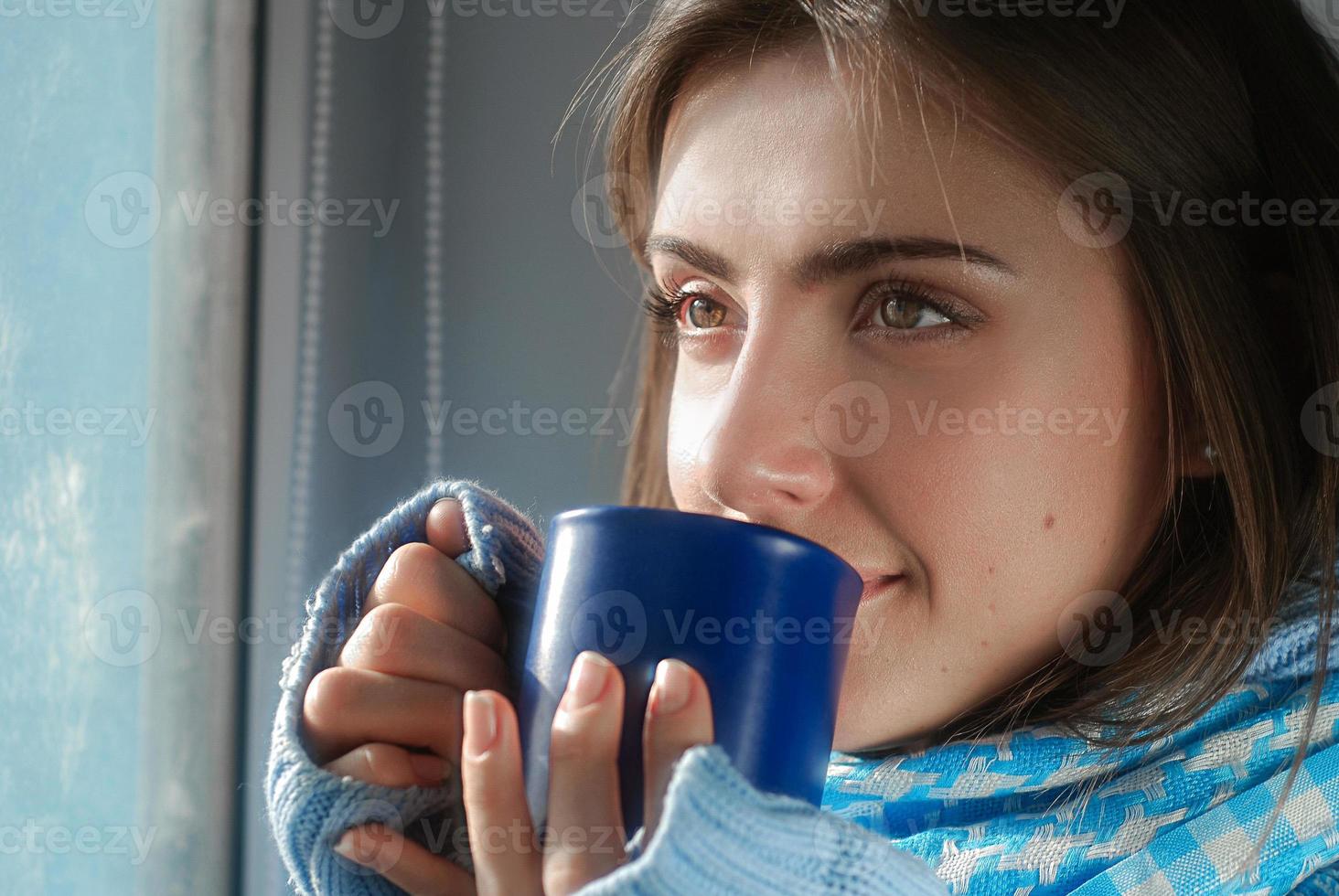 una mujer joven y triste que se queda en casa debido a la gripe y la temperatura en un suéter cálido y una bufanda con una taza de té cerca de la ventana foto