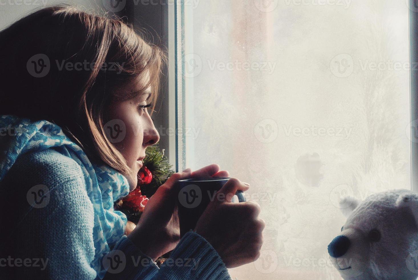 una mujer joven y triste que se queda en casa debido a la gripe y la temperatura en un suéter cálido y una bufanda con una taza de té cerca de la ventana foto